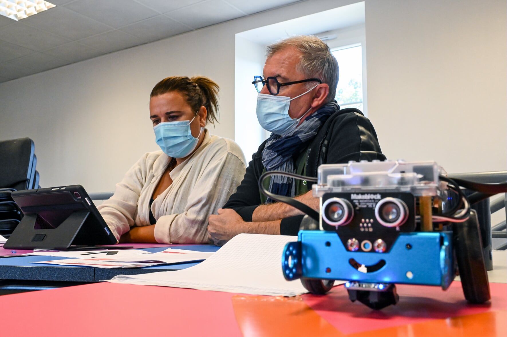 Deux personnes en formation robotique, travaillant sur une tablette avec un petit engin robotisé à côté.